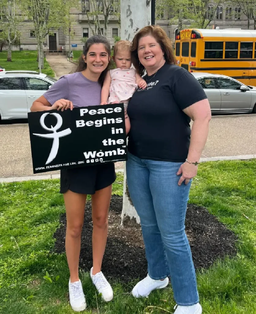 Two women holding a baby with a sign with the Feminists for Life logo that says Peace Begins in the Womb