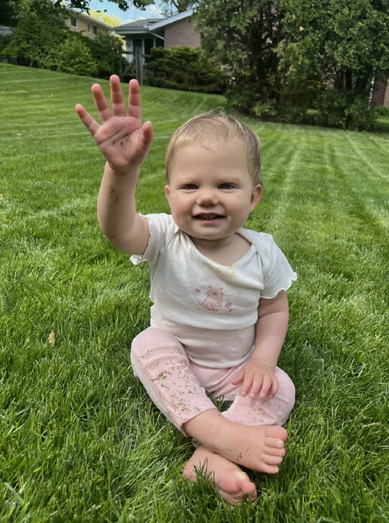 A toddler sitting in the grass and waving
